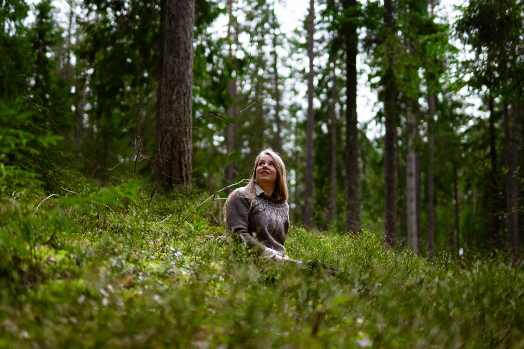 Pitkähiuksinen henkilö istuu vihreiden sammalten keskellä metsässä.
